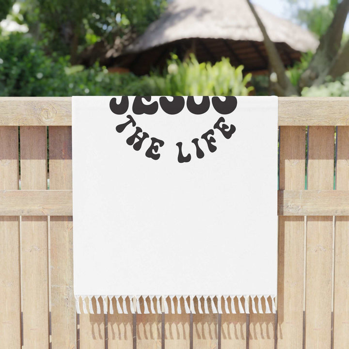 White Jesus Beach Cloth with black "Jesus The Life" text and knotted tassels hanging on wooden fence outdoors