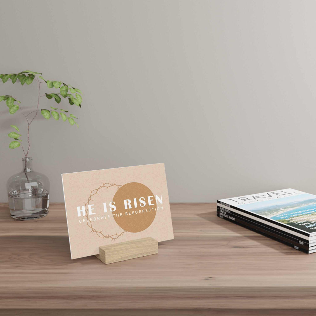Risen Gallery Board with hand-crafted oak stand displaying "He is Risen" art on desk with books and plant.