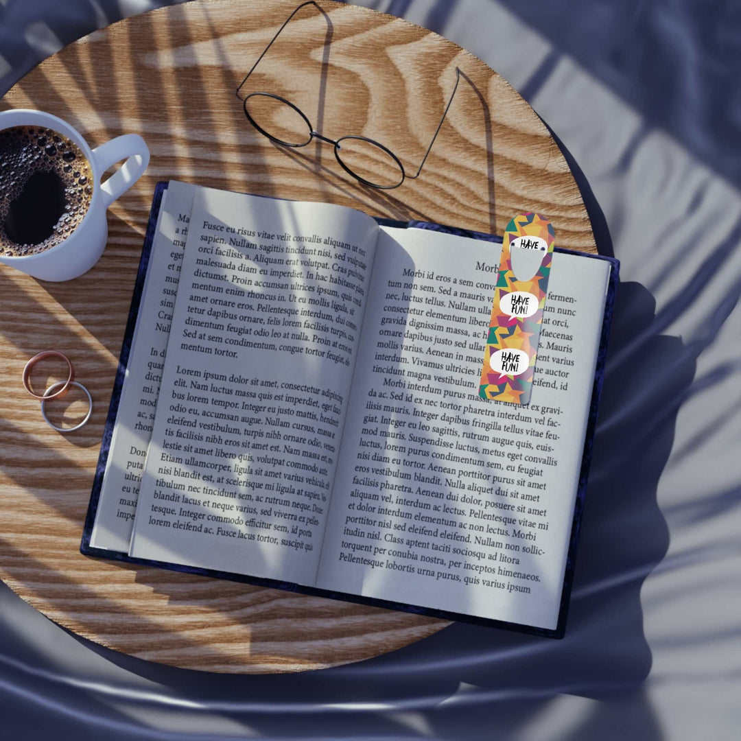 Have Fun Bookmark with colorful design on open book page, placed on wooden table next to coffee cup and glasses