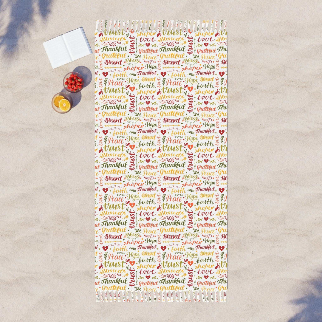 Christian Beach Cloth beach towel with colorful faith-inspired words and knotted tassels on sand next to an open book and drinks