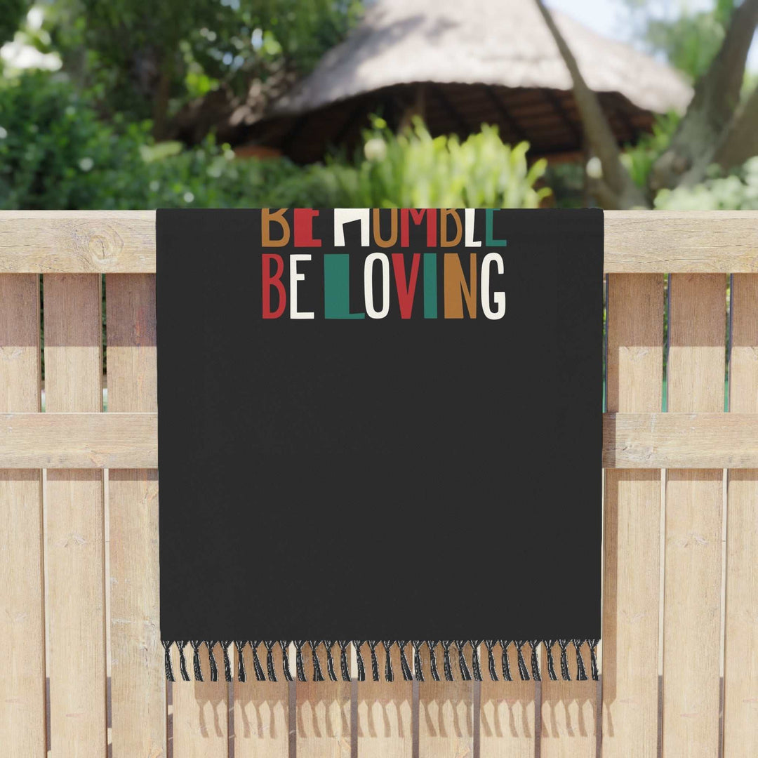 Bible Verse Beach Cloth with colorful "Be Humble Be Loving" text and knotted tassels hanging on wooden fence outdoors