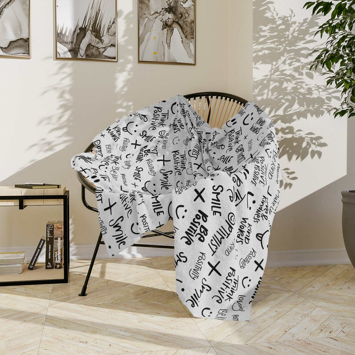 Soft white fleece Think Positive Blanket with black motivational words and symbols draped over a modern black chair in a sunny room