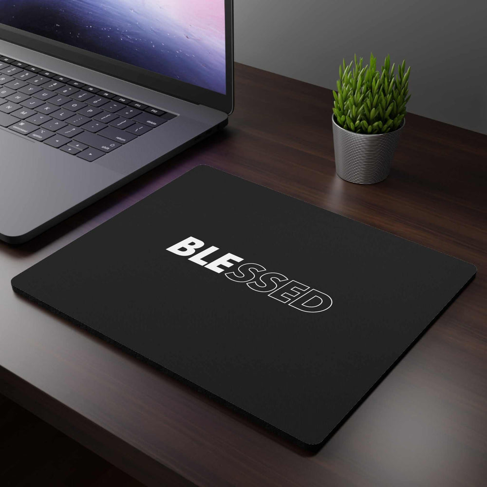 Black mouse pad with the word "BLESSED" in white letters on a wooden desk next to a laptop and small potted plant
