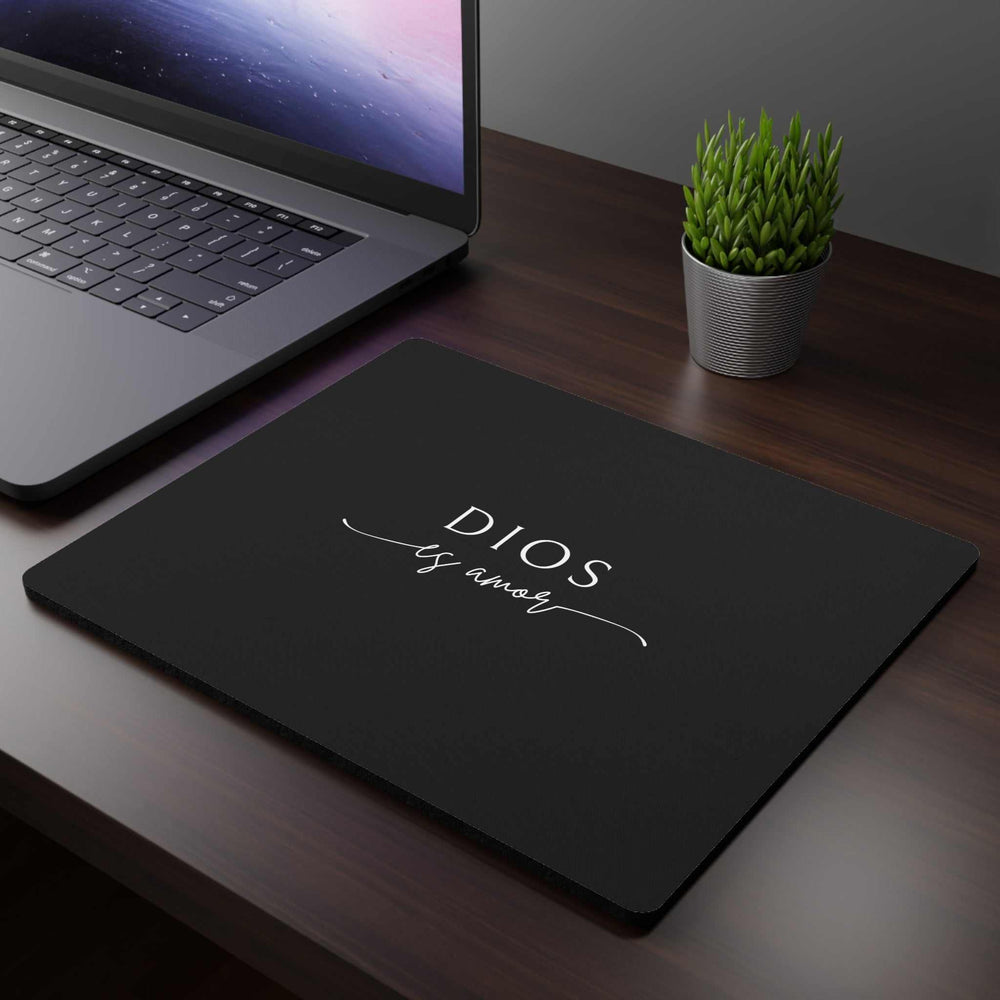 Black Dios Es Amor mouse pad with white script text on wooden desk next to laptop and small potted plant