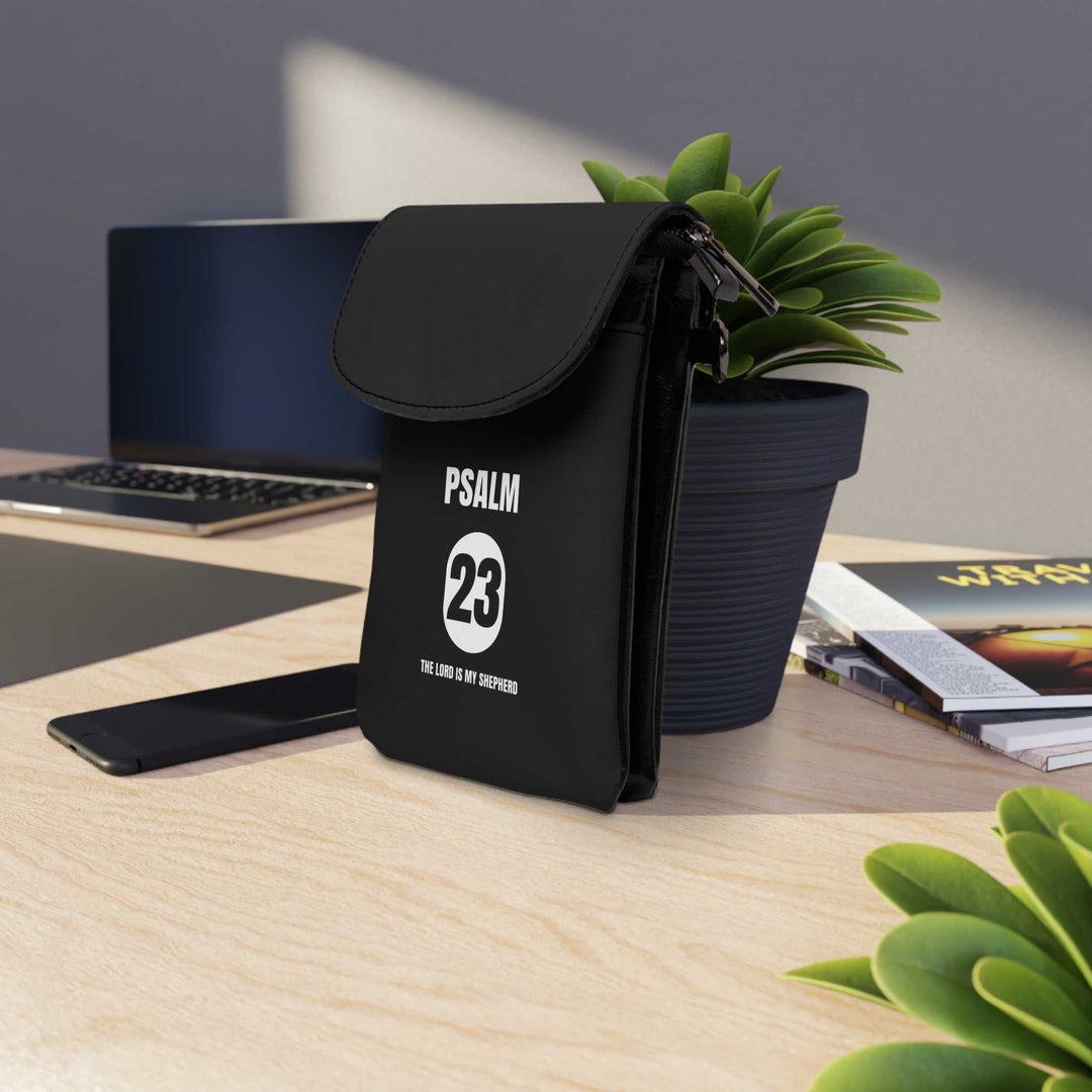 Black Psalm 23 Cell Phone Wallet with faux leather material and adjustable strap on wooden desk near laptop and plants