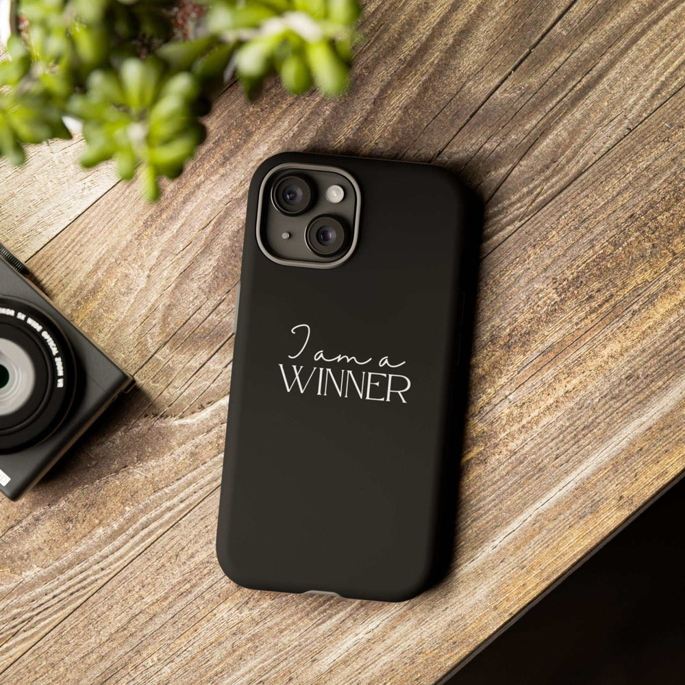 Black "Winner Mindset" phone case with matte finish, dual-layer protection, and motivational text "I am a WINNER" on wooden surface next to a camera.