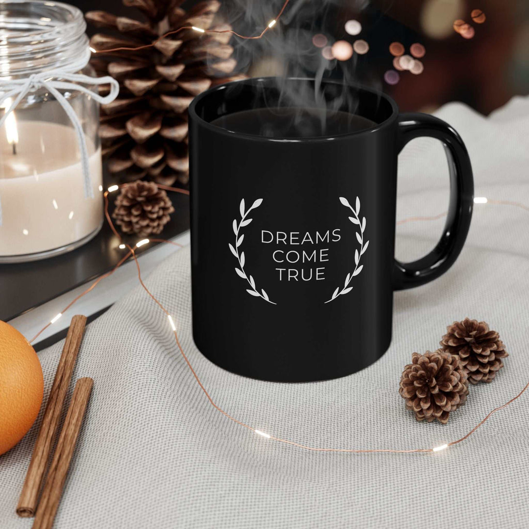 Black ceramic mug with "Dreams Come True" text and laurel design, steam rising, surrounded by pine cones and cinnamon sticks.