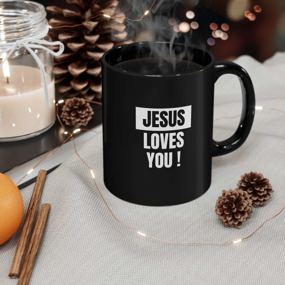 Black ceramic mug with "Jesus Loves You!" text, steam rising, surrounded by pine cones and candle on table