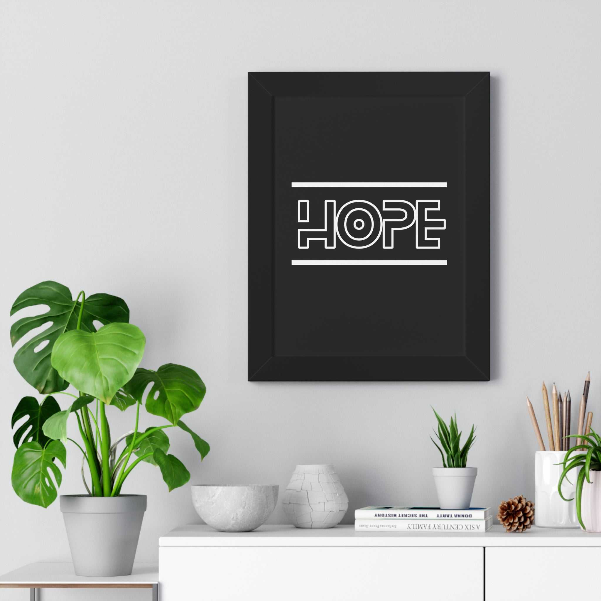 Black framed poster with the word "HOPE" in white modern typography hanging on a light gray wall above a white table with green plants and decorative objects