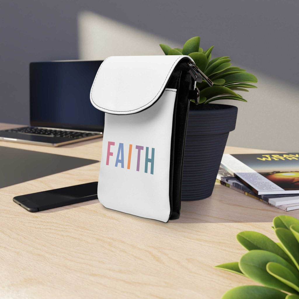 Faith-themed cell phone wallet in white faux leather with detachable strap and card slots placed on a desk near plants and laptop