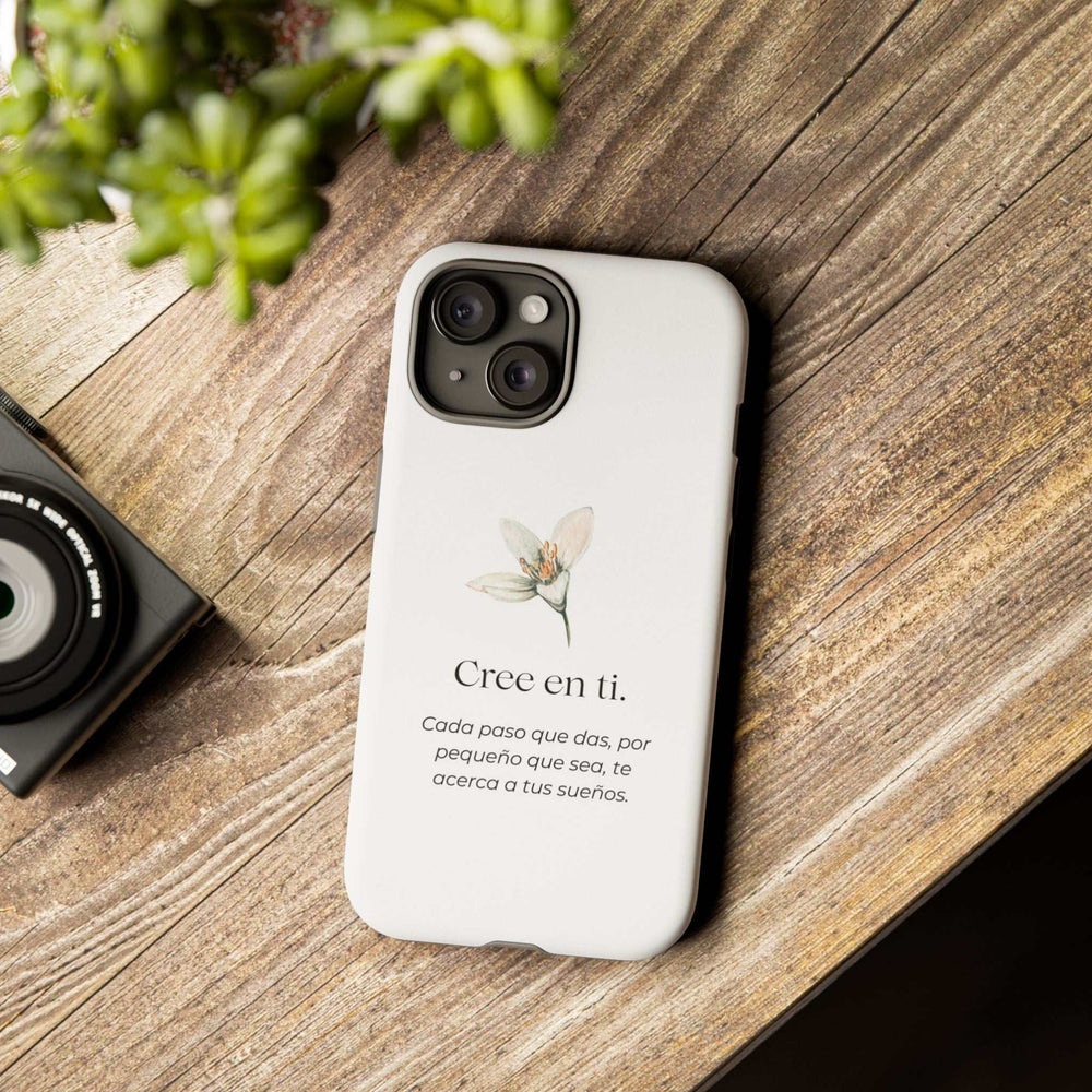 White Cree en Ti phone case with motivational Spanish text and floral design on wooden surface next to a camera, durable protective dual-layer with matte finish.
