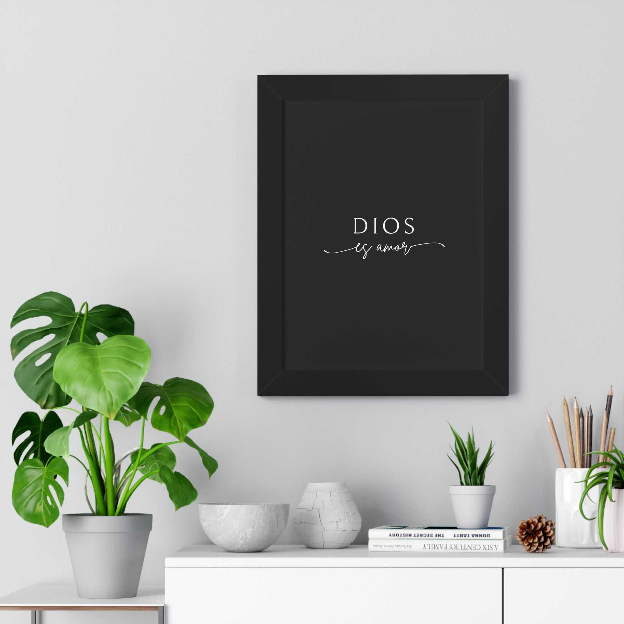 Black framed Dios Es Amor poster on white wall above white cabinet with plants and decor