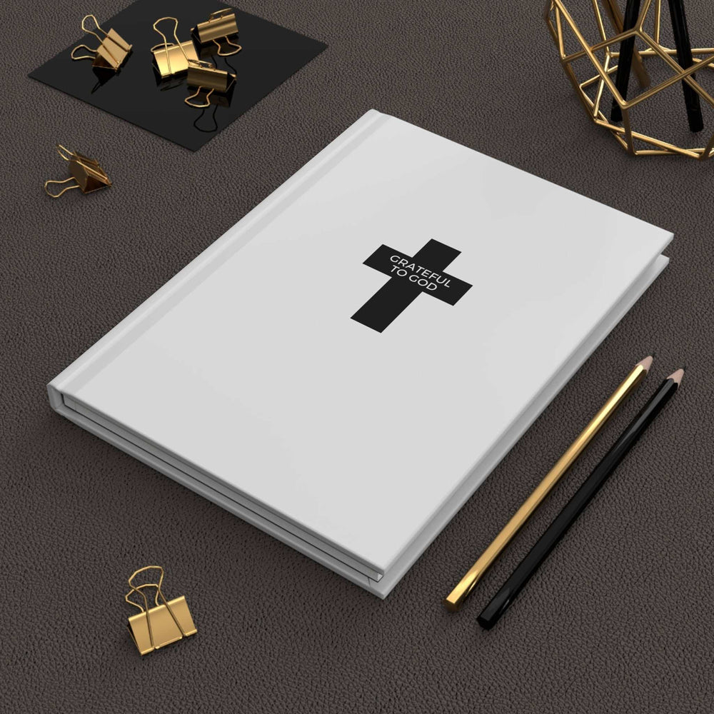 White hardcover journal with "Grateful to God" text and cross design, matte finish, surrounded by pencils and gold binder clips on dark textured surface