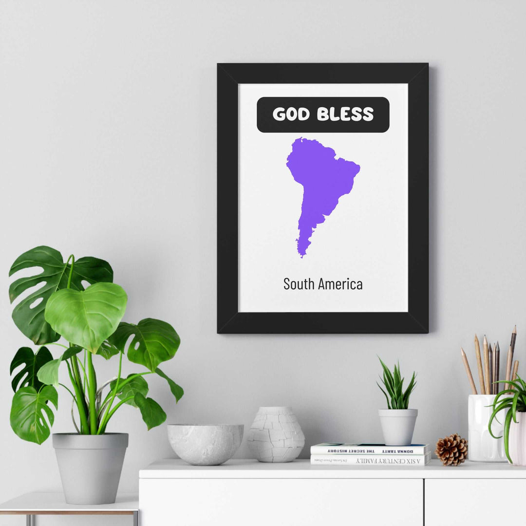 Framed God Bless South America poster with purple map and black text on white matte paper displayed on wall above modern shelf with plants and decor