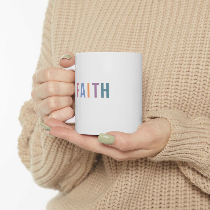 White ceramic mug with the word "FAITH" in colorful letters, held by hands with green nails, perfect for coffee or tea.
