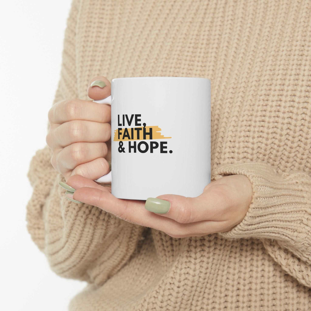 White ceramic mug with Live Faith & Hope text held by hands against beige sweater background