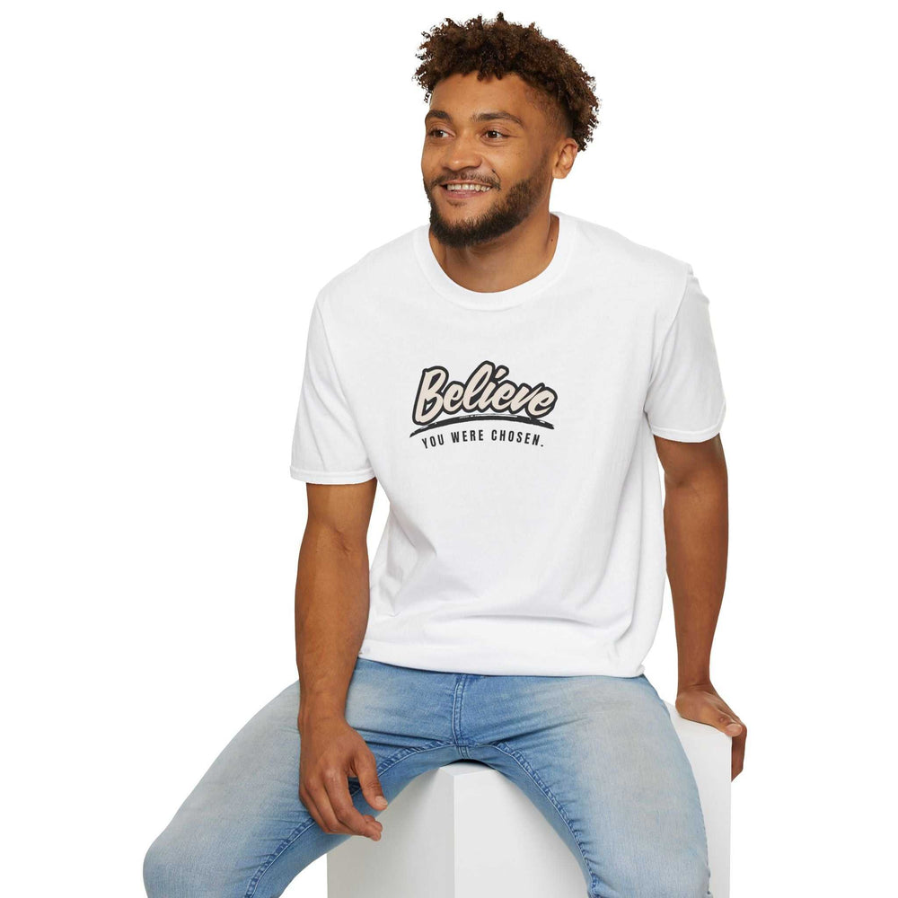 Man wearing white "Chosen & Beloved" T-shirt with "Believe you were chosen" text and light blue jeans sitting on a white cube