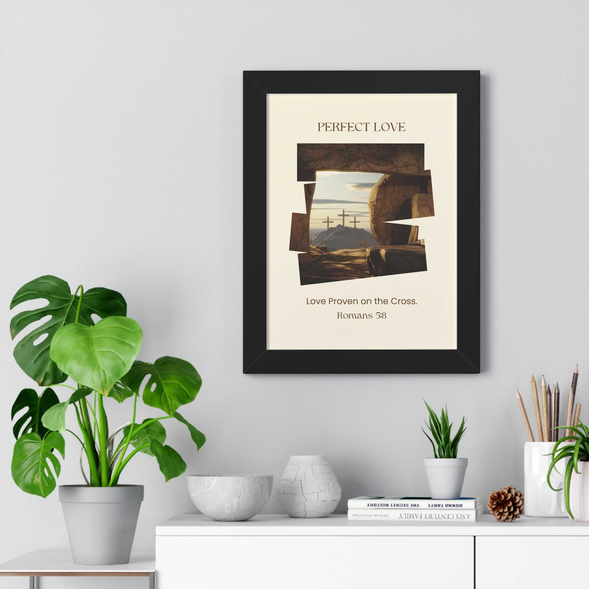 Framed Perfect Love poster with Christian cross and Romans 5:8 scripture on wall above white shelf with plants and decor