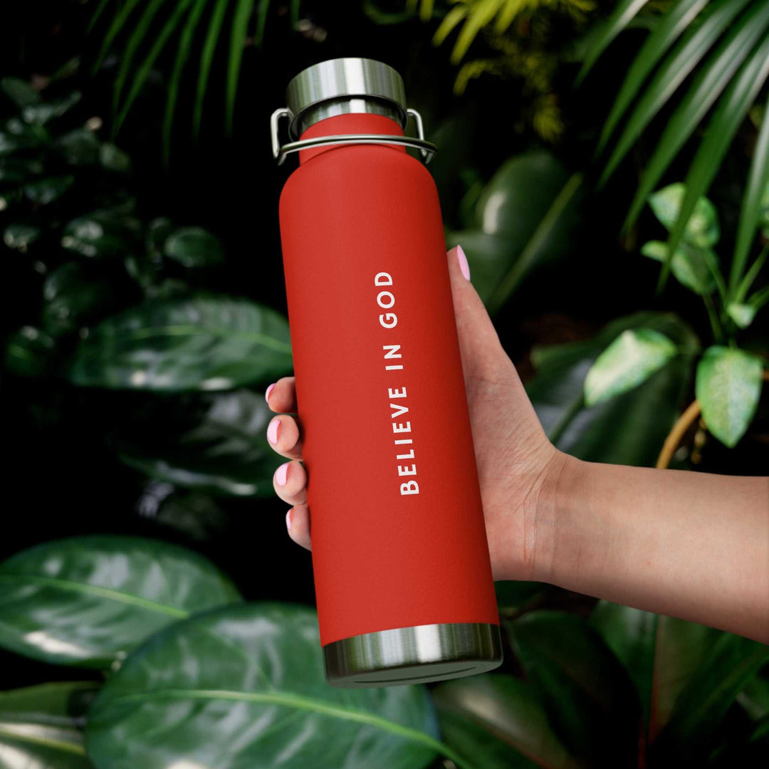 Red Believe in God bottle with stainless steel lid held against green leafy background