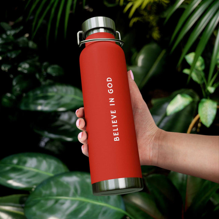 Red Believe in God bottle with stainless steel lid held against green leafy background