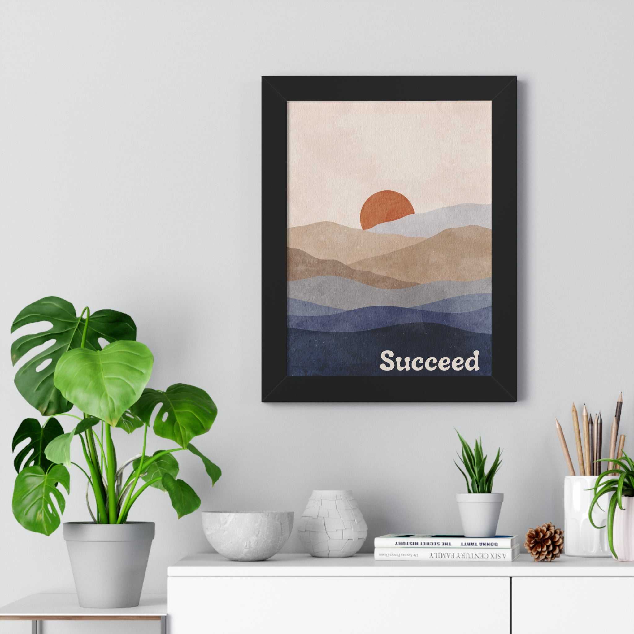 Succeed framed poster with motivational text and abstract landscape in black wood frame on wall above white cabinet with plants and decor