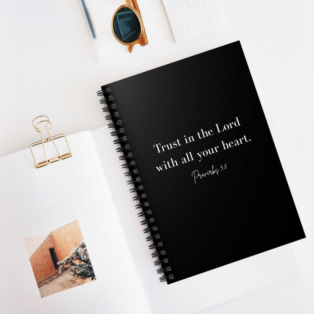 Black spiral-bound notebook with "Trust in the Lord with all your heart. Proverbs 3:5" printed on the cover, placed on a white desk next to an open book and sunglasses.