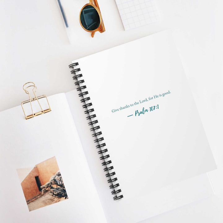 White spiral-bound notebook with Psalm 107:1 printed on the front cover, placed on a white desk with glasses and other office items.