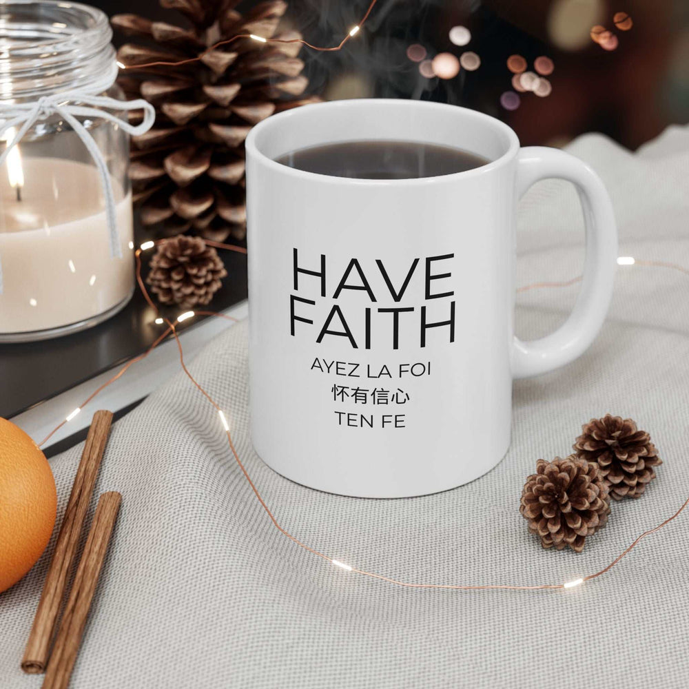 White ceramic "Have Faith" mug with black text in multiple languages, placed among pine cones and a candle on a cozy surface
