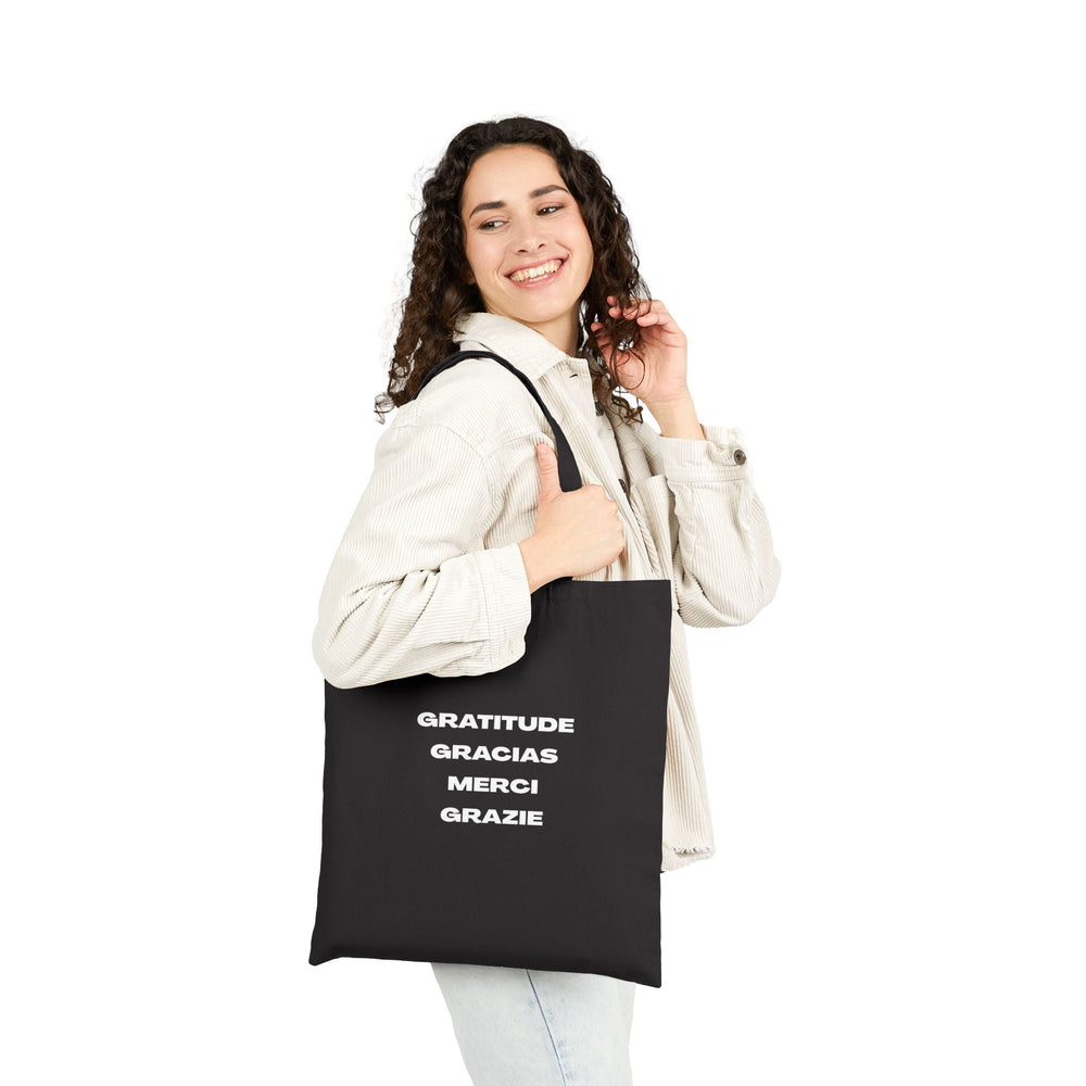 Black cotton canvas tote bag with gratitude text in multiple languages, carried by smiling woman in light jacket