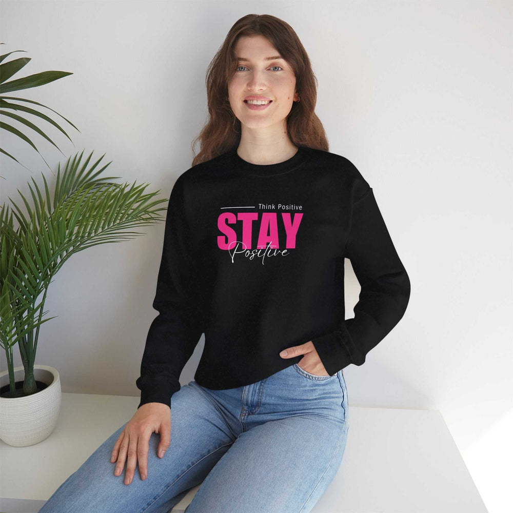 Woman wearing black Stay Positive sweatshirt with pink and white text, paired with light blue jeans sitting indoors near green plant