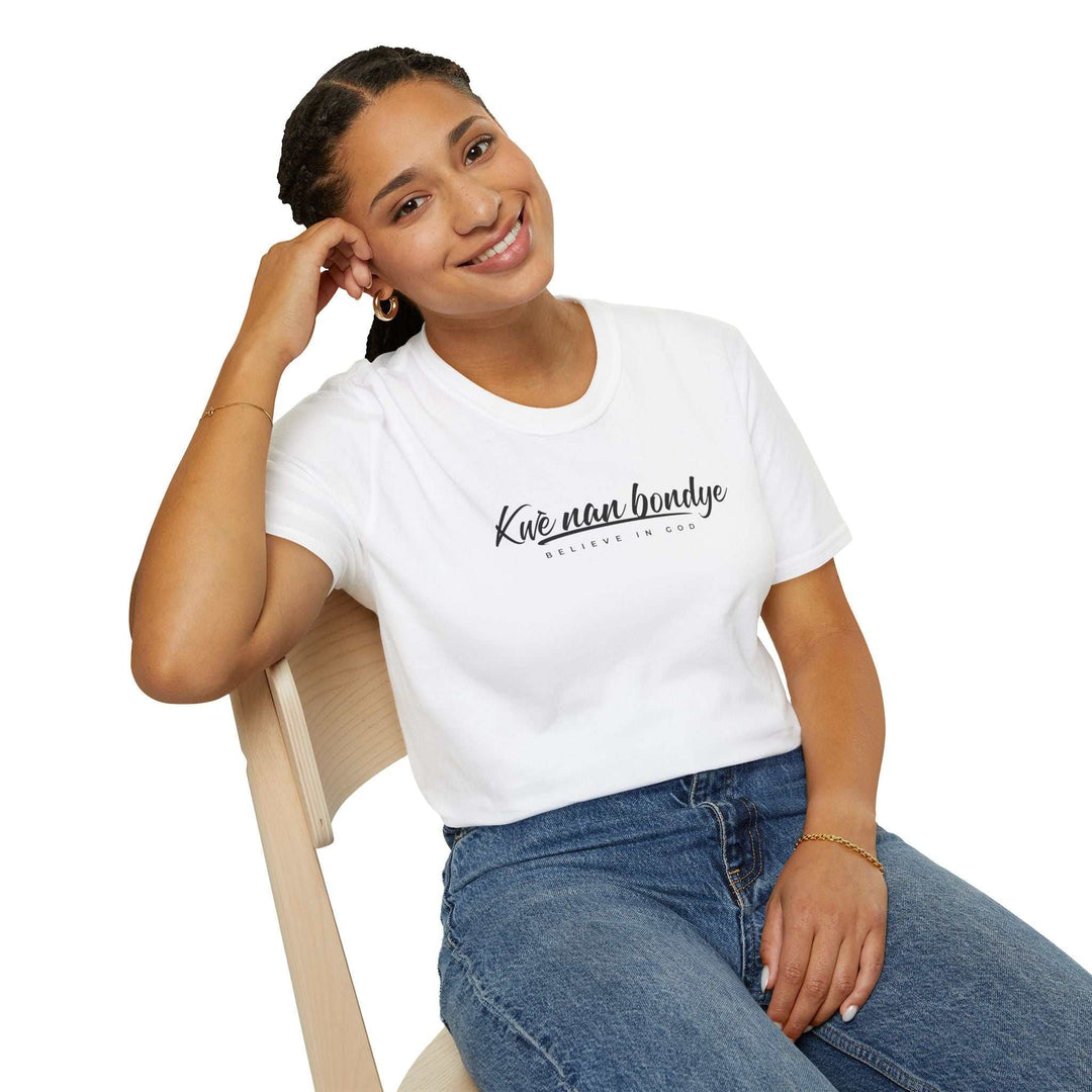 White Believe in God T-Shirt in Haitian Creole worn by smiling woman sitting on chair, paired with blue jeans